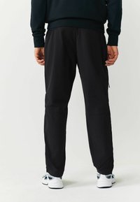 Schwarze Sporthosen mit entspanntem Schnitt, schmal zulaufenden Beinen und Reißverschlusstaschen an den Seiten. Getragen mit weißen und marineblauen Turnschuhen. Glatte Textur.