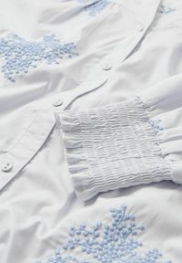 Camicia bianca in cotone con delicati ricami floreali azzurri e polsini arricciati per una vestibilità textured. Bottoni fissano la parte anteriore.