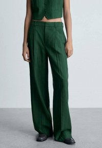 Pantalons larges verts à rayures verticales, avec une taille plate, des passants de ceinture et des poches latérales. Fabriqués en tissu lisse.
