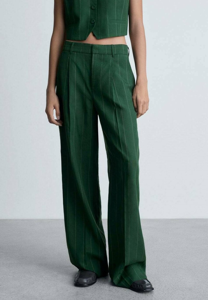 Pantalons larges verts à rayures verticales, avec une taille plate, des passants de ceinture et des poches latérales. Fabriqués en tissu lisse.