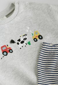 Grauer Baby-Pullover mit gesticktem Kuhmotiv, roten und gelben Traktoren sowie einem kleinen Stück grünem Gras, kombiniert mit navyblauen und weißen gestreiften Hosen.