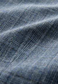 Gros plan d'un tissu bleu-gris texturé avec un motif de tissage visible et des plis légers créant de douces ombres.