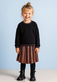 Schwarzer Pullover mit pinken Akzenten, metallisch plissierte bronzefarbene Rock, schwarze Strumpfhosen und klobige schwarze Stiefel. Kind lächelt vor einem hellblauen Hintergrund.