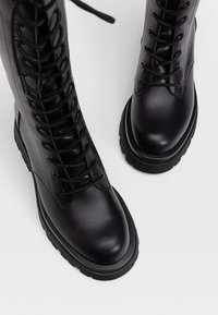 Botas de combate de cuero negro con cordones y suelas gruesas y acanaladas sobre un fondo blanco liso, vistas desde arriba.