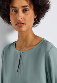 Blusa teal in tessuto satinato, con scollo rotondo, chiusura a 1 bottone e dettaglio a forma di chiave sul petto, caratterizzata da maniche lunghe e fluide.
