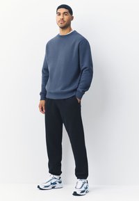 Sötétkék pulóver kerek nyakkivágással és bordázott mandzsettákkal, fekete melegítőnadrággal és kék részletekkel díszített fehér sportcipővel párosítva.