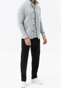 Chaqueta de sudadera gris claro con un cuello alto, botones frontales y bolsillos laterales, combinada con pantalones negros y zapatillas blancas.