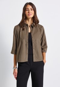 Chemise marron à boutons en tissu doux et texturé, avec un col et des manches retroussées. Portée par-dessus un débardeur noir.