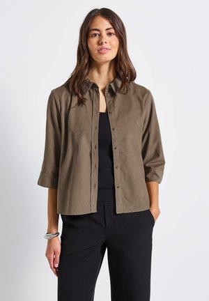 Camicia marrone con bottoni realizzata in un tessuto morbido e texturizzato, con colletto e maniche arrotolate. Indossata sopra un top nero.