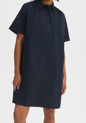 Woman wearing a loose-fitting, short-sleeve, navy blue shirt dress with a collar and three buttons on a plain background.