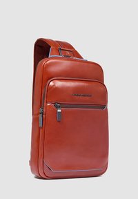 Mochila de cuero marrón con textura suave, que cuenta con un bolsillo frontal con cremallera y detalles en azul, diseño estructurado y correa ajustable.