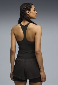 Γυναίκα με πιασμένα πλεξούδες, φορώντας μαύρο αθλητικό τοπ χωρίς μανίκια με αγκώνα racerback και μαύρα στρωματοποιημένα αθλητικά σορτς, στέκεται μπροστά σε γκρι διαβαθμισμένο φόντο.