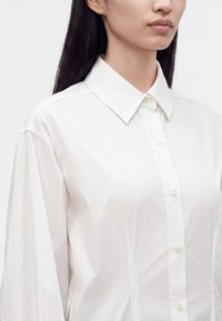 Femme aux longs cheveux foncés portant une chemise blanche à boutons avec un col pointu, vue des épaules jusqu'au milieu de la poitrine sur un fond uni.