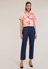Blusa rosa a maniche corte con motivi tie-dye rossi e bottoni, abbinata a jeans blu a gamba dritta e décolleté borgogna.