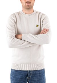 Pull en tricot beige clair avec poignets et ourlet côtelés, présentant un petit logo jaune brodé sur la poitrine. Conception standard avec col rond.