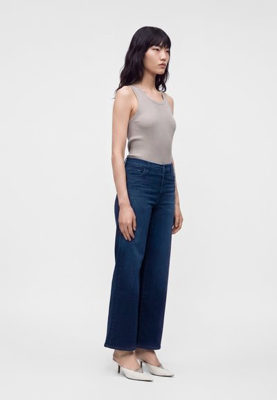 Femme aux longs cheveux foncés portant un haut beige sans manches, un jean large bleu foncé et des chaussures blanches à talons hauts, debout de côté.