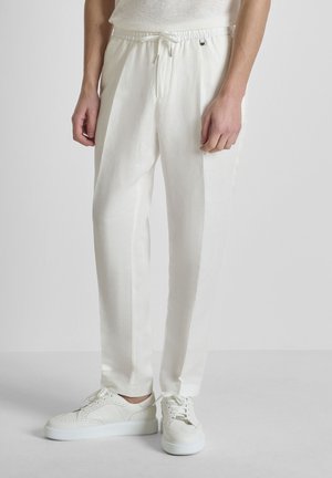 Muškarac u bijelim hlačama s vezicama i bijelim tenisicama, stoji na jednostavnoj svijetloj pozadini.