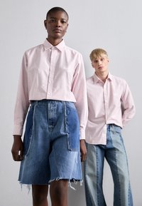 Rutet rosa skjorte med krage, kombinert med slitte blå denimshorts. Modellen står mot en nøytral bakgrunn.