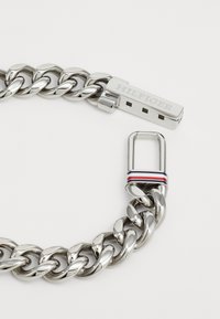 Masivna srebrna narukvica od poliranog metala s pravokutnom kopčom na kojoj je ugravirano "HILFIGER" i prugastim detaljem u crvenoj, bijeloj i plavoj boji.