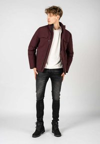 Giubbotto imbottito borgogna con bottoni a pressione e due tasche sul petto, indossato sopra una camicia bianca, abbinato a jeans scuri e stivali neri stringati.