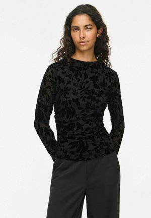 Femme aux longs cheveux bouclés portant un haut noir à manches longues avec motif floral et un pantalon noir, debout les mains derrière le dos.