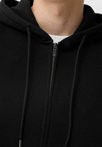 Detailní záběr na osobu v černé mikině na zip s tkaničkami přes bílou košili, se zaměřením na výstřih a zip.