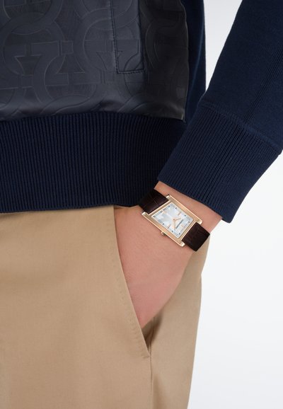 Χέρι με ορθογώνιο χρυσό ρολόι με λευκό καντράν και καφέ δερμάτινο λουράκι, χέρι στην τσέπη μπεζ παντελονιού, ορατή μανσέτα μπλε ριγωτού πουλόβερ.