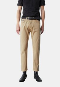 Pantaloni in cotone beige con vestibilità dritta e leggermente affusolata. Presentano tasche frontali e una cintura. Abbinati a stivaletti neri alla caviglia.