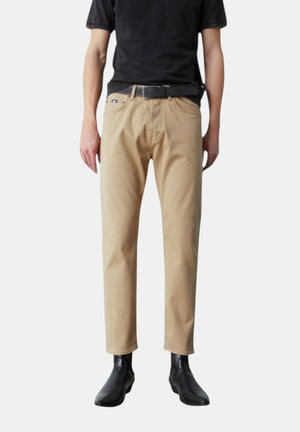 Pantaloni in cotone beige con vestibilità dritta e leggermente affusolata. Presentano tasche frontali e una cintura. Abbinati a stivaletti neri alla caviglia.