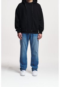Sudadera negra con cremallera y bolsillos frontales, combinada con jeans azul claro. Zapatillas blancas completan el look casual.