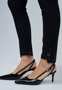 Tacones slingback negros con puntera puntiaguda y acabado brillante, con tiras finas y un tacón stiletto bajo. Llevados con jeans skinny oscuros con detalles en los dobladillos.