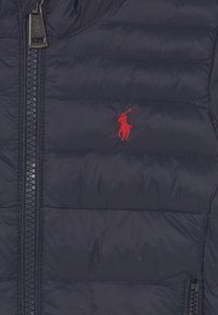 Tmavě modrá péřová bunda z nylonu, vybavená celým zipem a vodorovnými švy. Na hrudi je výrazné červené logo polo.