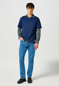 T-shirt navy sovrapposto a una camicia a quadri verde e blu, abbinato a jeans in denim azzurro chiaro e scarpe nere. Design semplice, vestibilità casual.