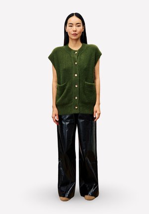 Femme debout portant un gilet en tricot sans manches vert, avec des boutons et des poches, et un pantalon en cuir noir à jambes larges, devant un fond uni.