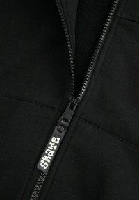 Czarna kurtka zapinana na zamek błyskawiczny z gładką teksturą materiału oraz wyraźnym zamkiem. Uchwyt zamka ma biały nadruk z logo.