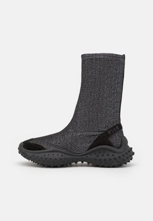 Bottine sneaker noire montante avec semelle texturée et dessus en maille, présentant un talon légèrement surélevé et un détail du nom de la marque à l'arrière.