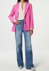 Blazer in velluto a coste rosa con rever a punta, abbinato a jeans a gamba larga blu chiaro. L'outfit include una camicia bianca con bottoni e scarpe con fantasia.