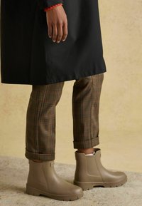 Botoși de gleznă impermeabili de culoare maro cu o talpă texturată, purtați cu pantaloni cadrilati răsuciți la manșete și o haină neagră parțial vizibilă.