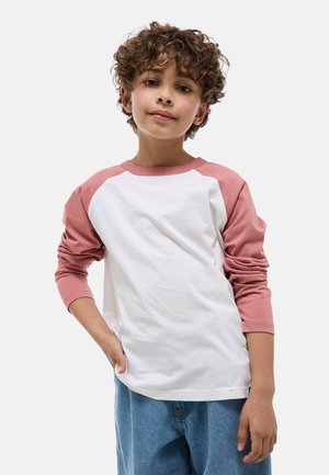 Garçon aux cheveux bouclés portant une chemise blanche à manches raglan roses et un jean bleu, debout avec une main dans la poche.