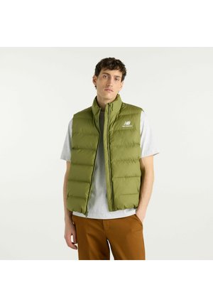 Giovane uomo che indossa un gilet imbottito verde oliva, una t-shirt grigia e pantaloni marroni, che posa con una mano in tasca su uno sfondo semplice.