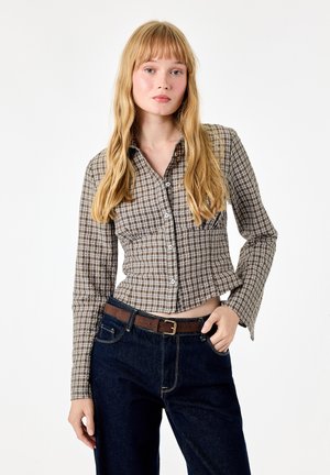 Jeune femme aux longs cheveux blonds portant une chemise à carreaux marron boutonnée et un jean bleu foncé avec une ceinture marron, posant avec une main dans la poche.