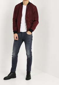 Burgundy bomberjacka med svarta ribbade muddar och krage, kombinerad med mörka denimjeans och svarta snörkängor. Ren, slät tygstruktur.