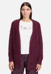 Kobieta nosząca puszysty, ciemnofioletowy kardigan na białej koszuli z ilustracjami butelek perfum oraz pasującymi, ciemnofioletowymi spodniami.