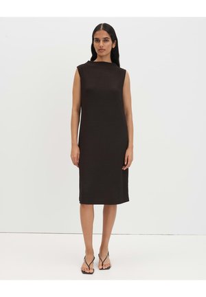 Vrouw staand, gekleed in een zwarte mouwloze knielange jurk en zwarte sandalen met bandjes en hak op een effen lichte achtergrond.