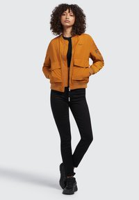 Oranger Bomberjacke mit zwei Fronttaschen, gerippten Bündchen und Reißverschluss vorne. Über einem schwarzen Oberteil getragen und kombiniert mit schwarzen Jeans und Turnschuhen.