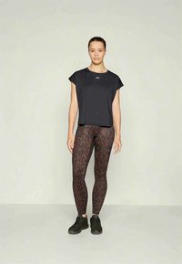 Naised seisab ettepoole, seljas must lühivarrukatega topp, pruun leopardimustriga legginsid ja mustad tossud, tasase tausta ees.