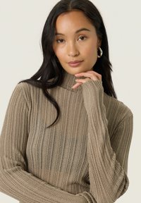Kvinde med langt mørkt hår, iført sølvfarvede hoop øreringe og en beige ribstrikket sweater, som hviler sin hånd forsigtigt nær sin hage.