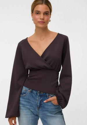 Jeune femme portant un haut noir à manches longues style cache-cœur et un jean bleu, debout avec une main dans la poche sur un fond uni.