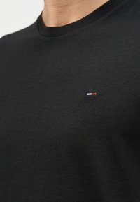 Κοντινό ενός ατόμου που φοράει μαύρο t-shirt με στρογγυλή λαιμόκοψη και μικρό ορθογώνιο λογότυπο σε κόκκινο, λευκό και μπλε στο στήθος.