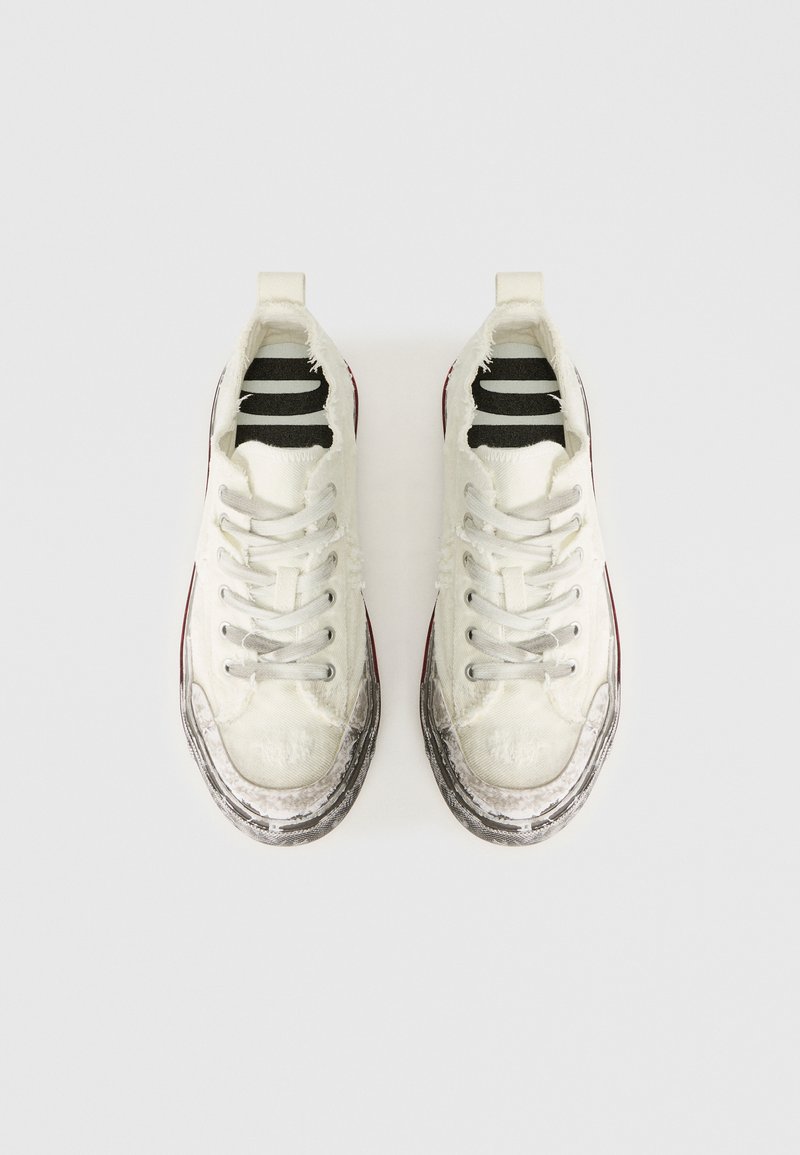 Zapatillas de lona blancas con bordes deshilachados, ojales plateados y suelas de goma texturizada. Se pueden ver plantillas a rayas en blanco y negro en el interior.